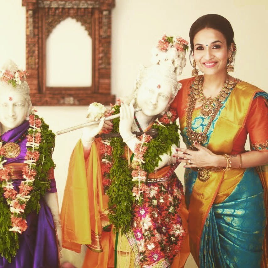 Soundarya Rajinikanth in an orange and turquoise silk saree standing beside garlanded temple figures